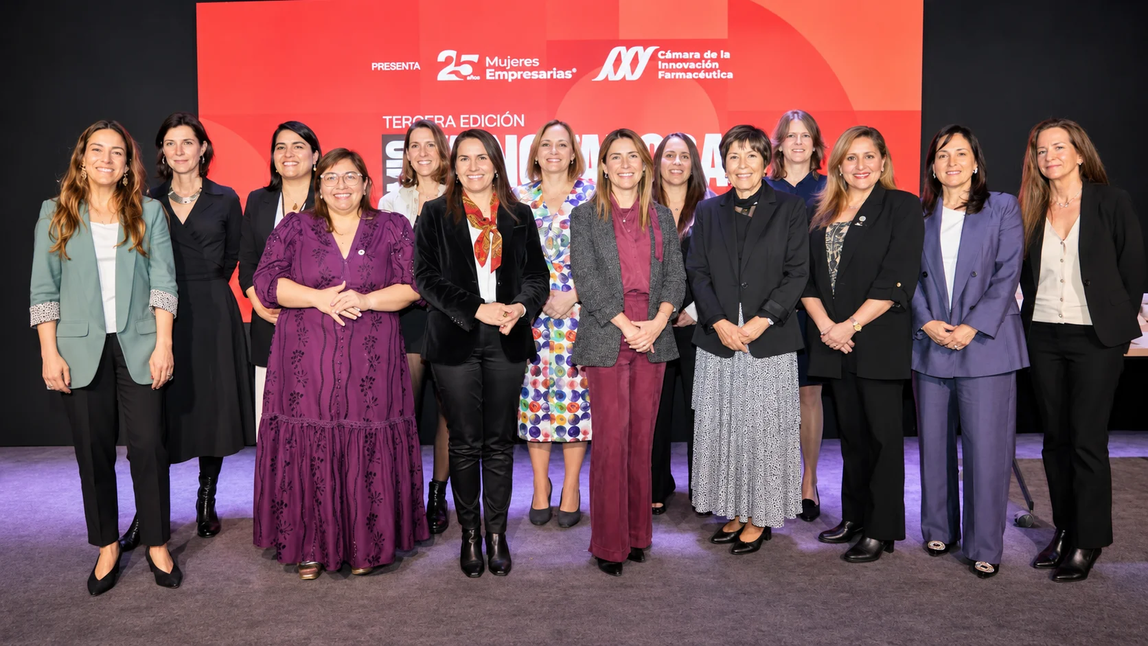 Grupo de mujeres posa sobre un escenario frente a una pantalla roja con la identidad del Premio Innovadoras en Salud 2026.